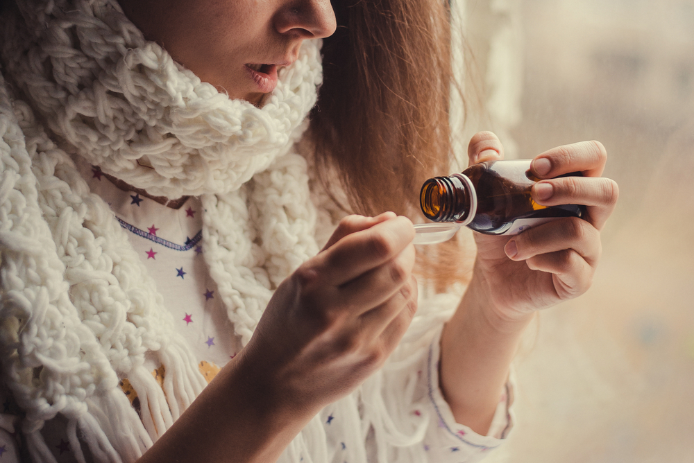 A woman with a polka dot shirt and a thick white scarf around her neck pours some cough medicine into a spoon. 