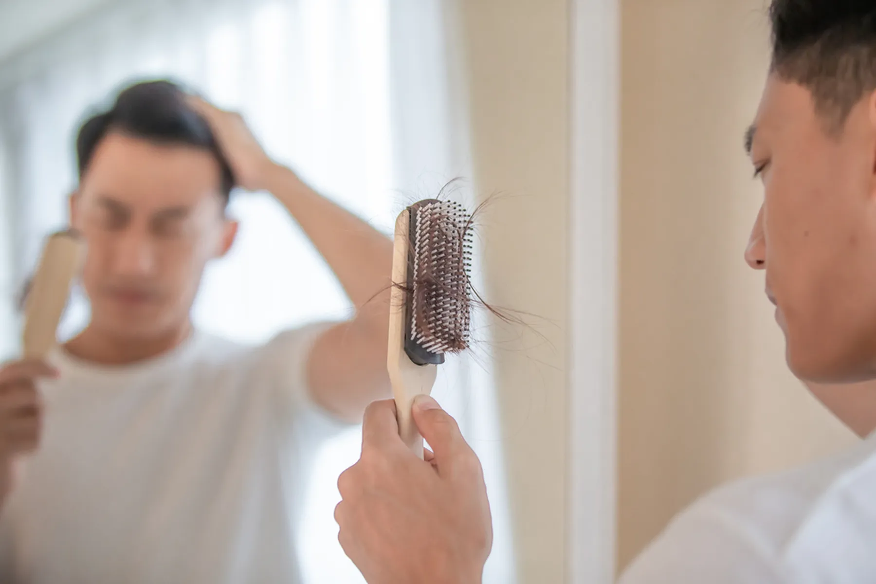A man looks at a hairbrush with that has a lot of his hair in the bristles. The man's reflection can be seen in the mirror.