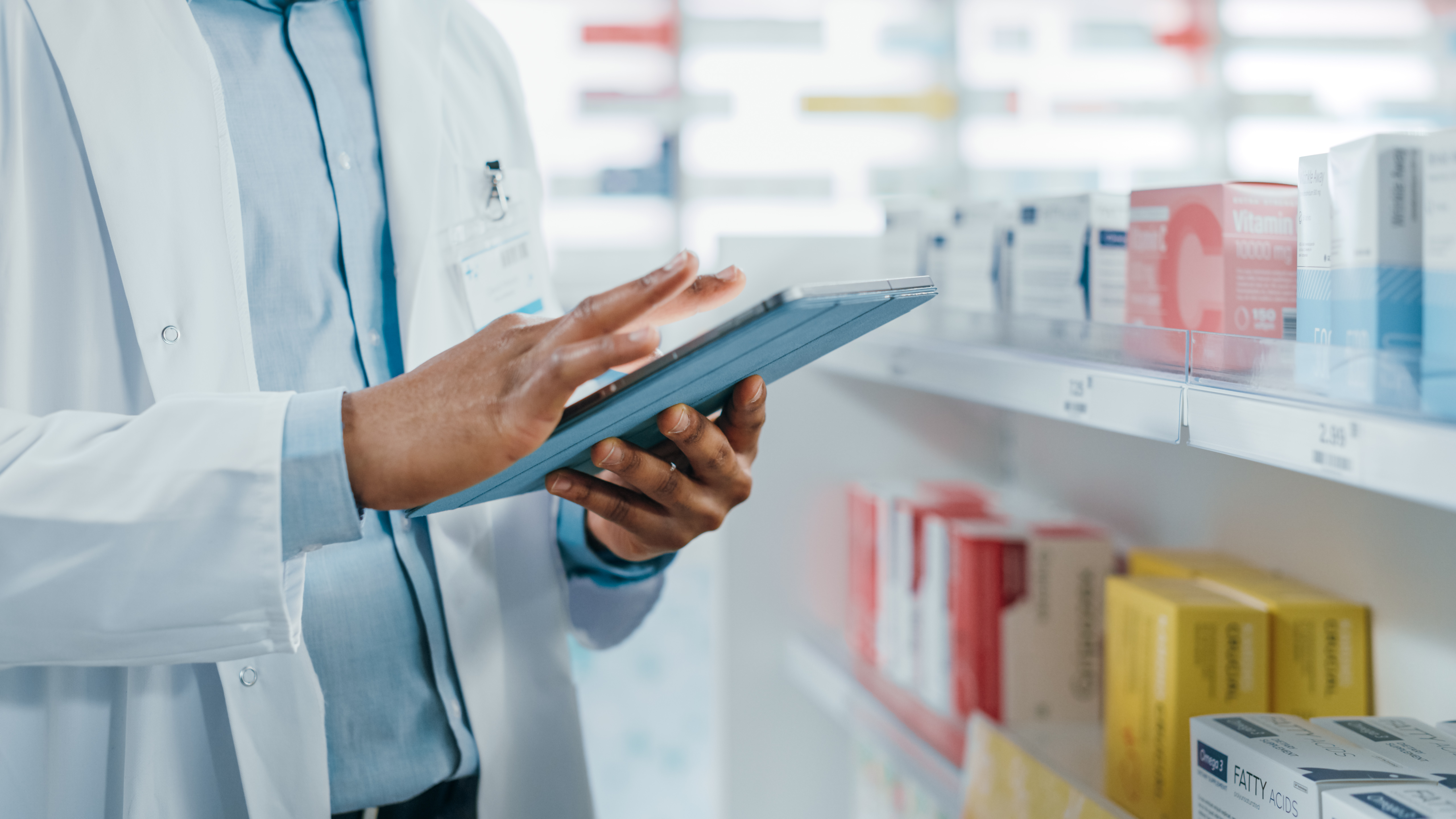 A pharmacist stands in front of shelves of medication and refers to a tablet.