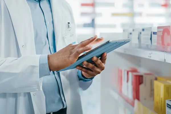 A pharmacist stands in front of shelves of medication and refers to a tablet.