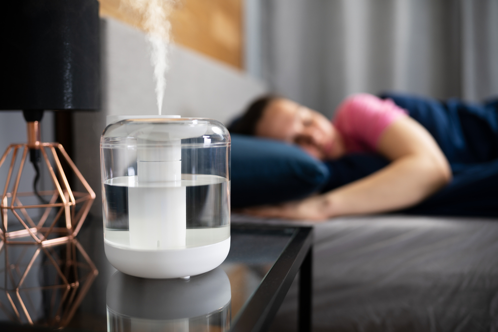 A woman in a pink shirt sleeps in bed as a humidifier runs next to her on her nightstand.