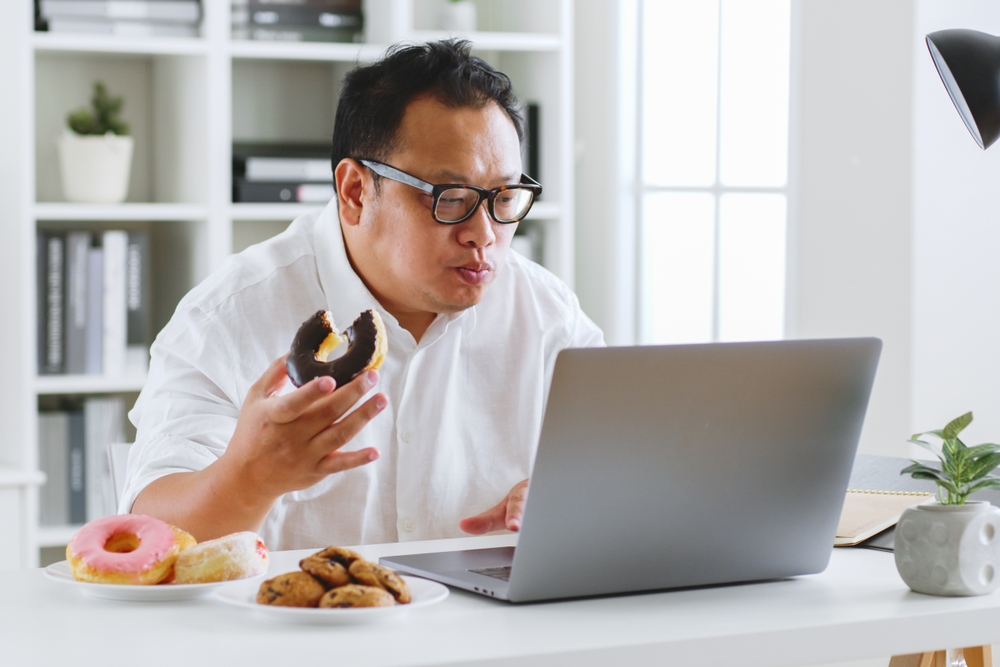 A man sits at his laptop and eats a chocolate donut, with a plate of donuts and cookies next to him.
