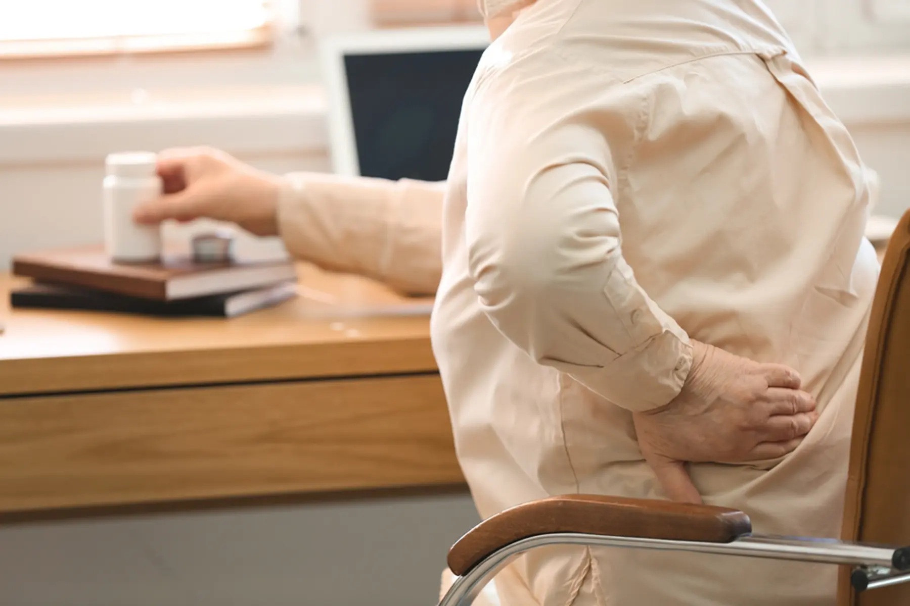 An adult in an eggshell-colored button-down shirt sits in an office chair. Their left hand is holding their mid-back, and their right hand is on a bottle of medication. 