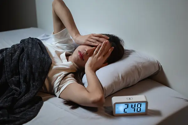 A woman lays in her bed with a clock reading 2:48am and holds her hands on her head.
