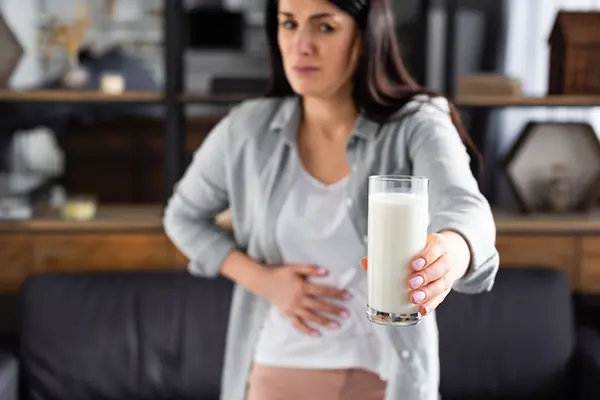 A woman looks at a glass of milk she is holding with a concerned and upset face, as she clutches her stomach with one hand.