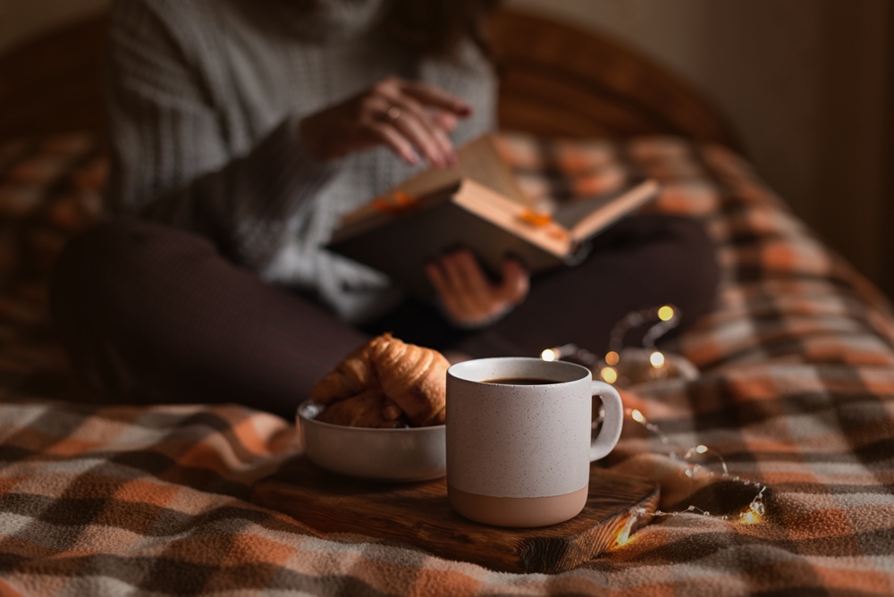 A person reading a book on a bed in a dark room with a cup of coffee along with a bowl containing a croissant. 