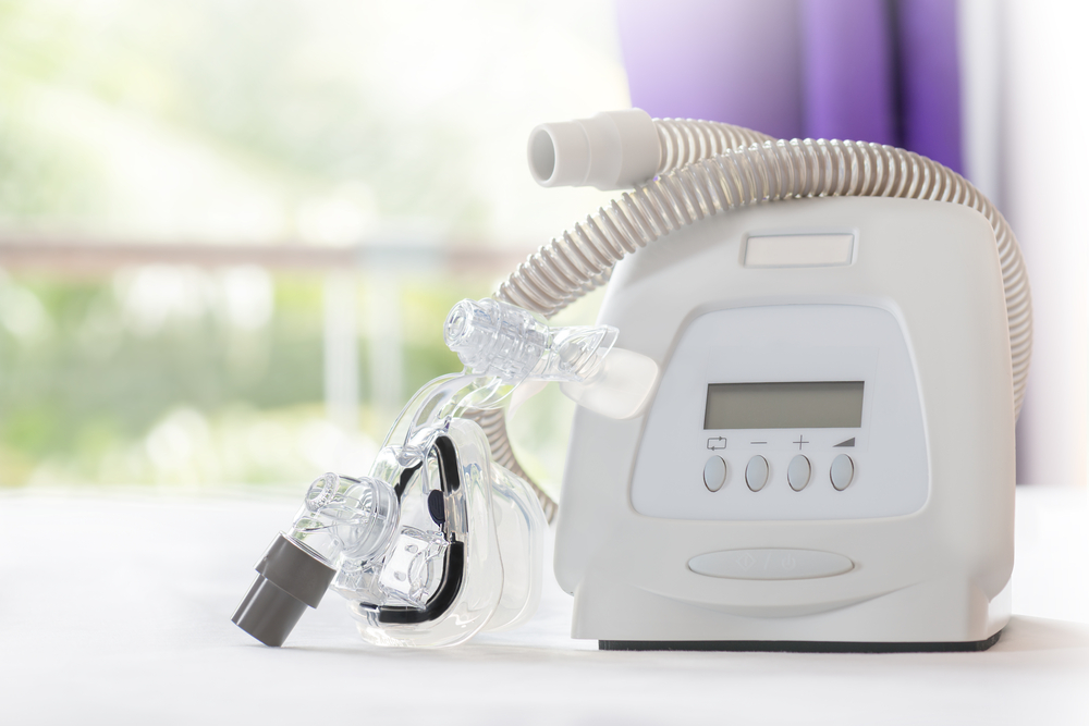 A CPAP machine on a white table in front of a window with a purple curtain. 