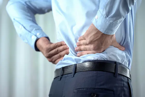 A man in a dress shirt and dress pants holding his lower back with both hands, likely because his back is hurting. 