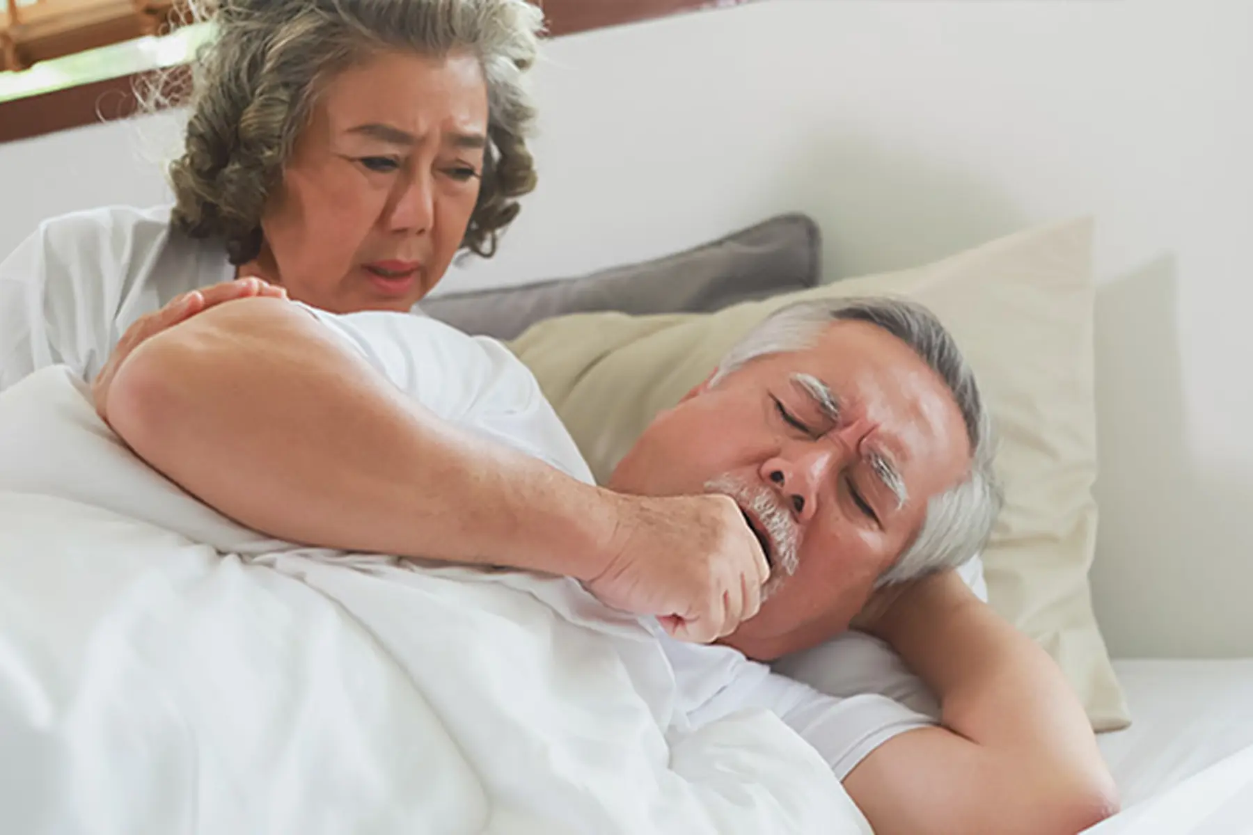Man with tonsillitis coughing in bed as his wife looks at him concerned.
