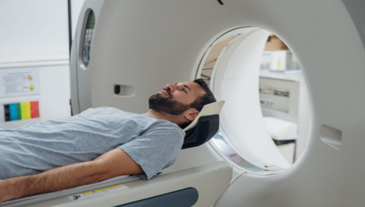 A man with dark hair and a dark beard lying down before going into a medical imaging machine.