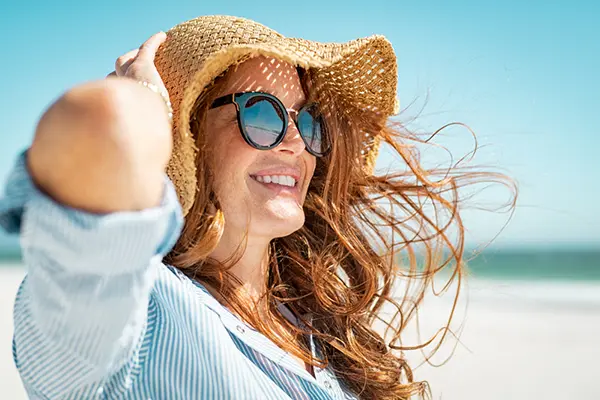 Woman at the beach wearing a shirt to cover her skin, sunglasses, and a hat.