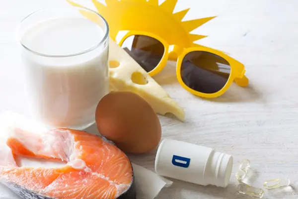 A piece of salmon, a glass of milk, an egg, cheese, and a bottle of vitamin D supplements on a table next to yellow sunglasses and a picture of the sun.