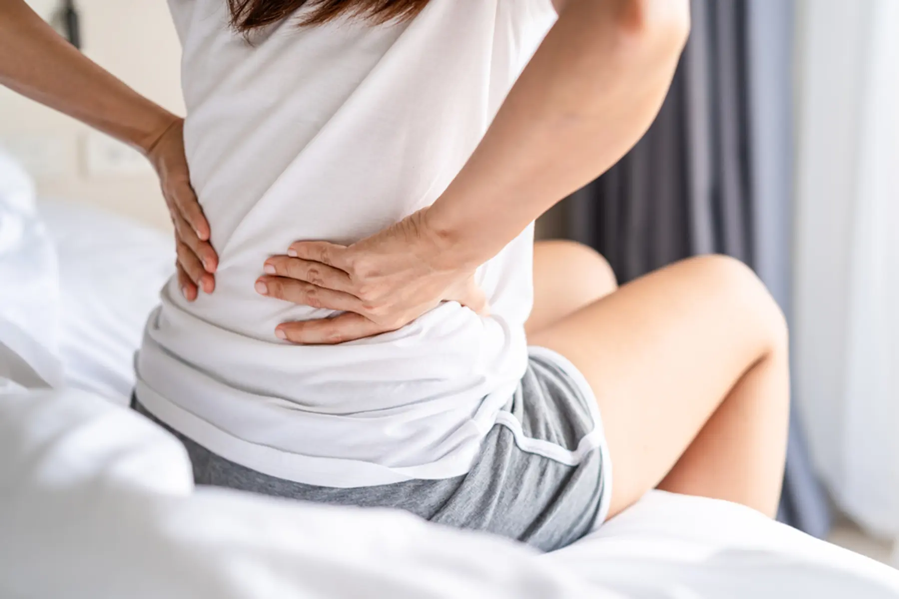 A woman in a white t-shirt and grey shorts sits up on the edge of the bed as she holds her hands on her mid to lower back.