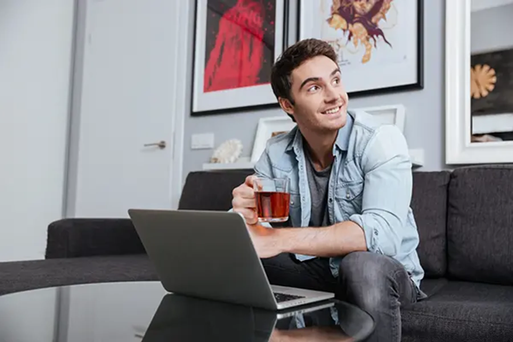 A man sits on a couch opposite a laptop and smiles while holding a cup of tea.