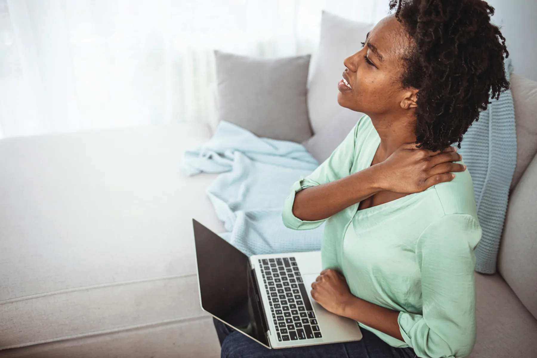 A woman with a laptop on her lap holding her neck/shoulders. She appears to be physically uncomfortable. 