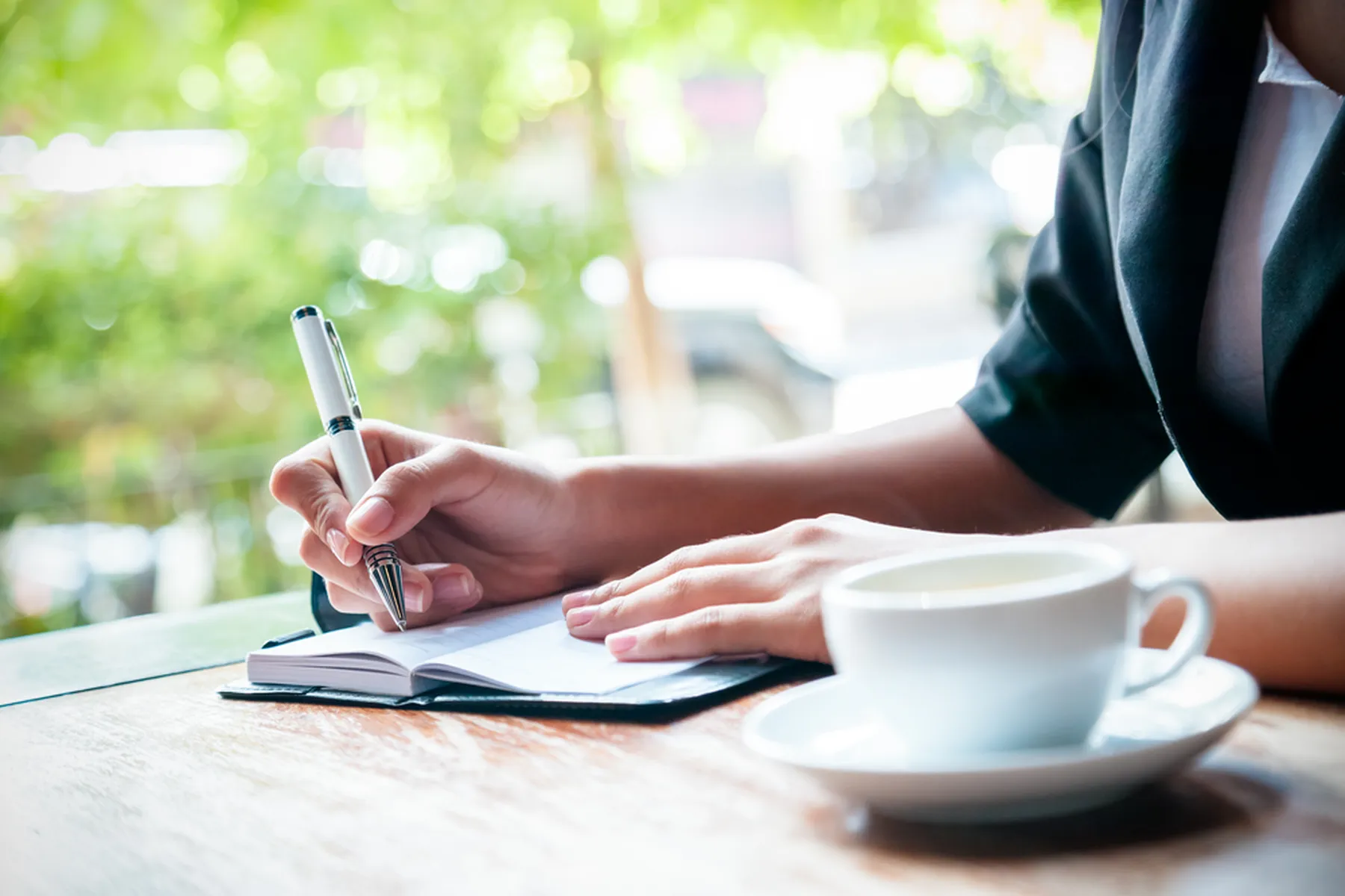 A person's hands are seen writing in a journal with a coffee cup next to them.