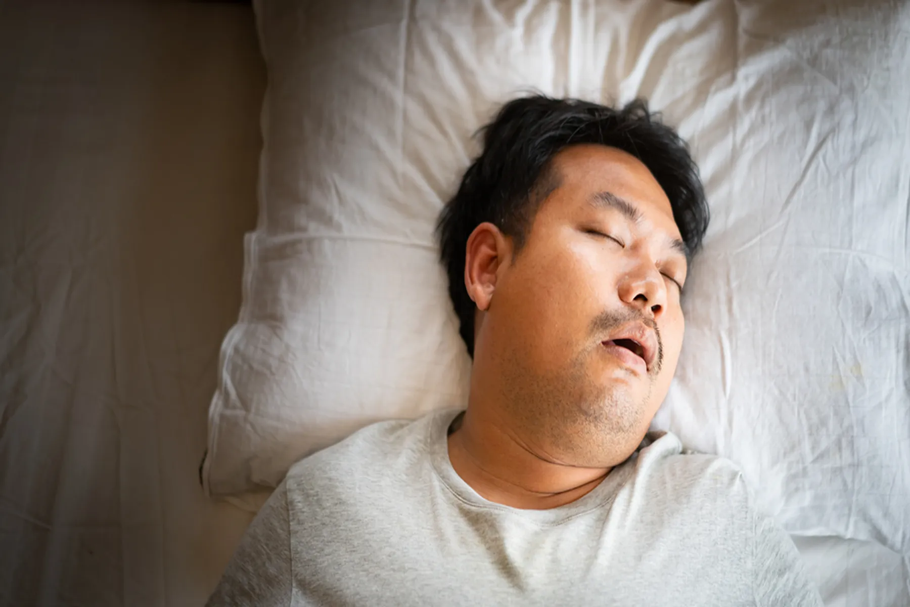  A young man in a white t-shirt lies on a white pillow. He is sleeping with his mouth partly open, and he looks to be snoring. 