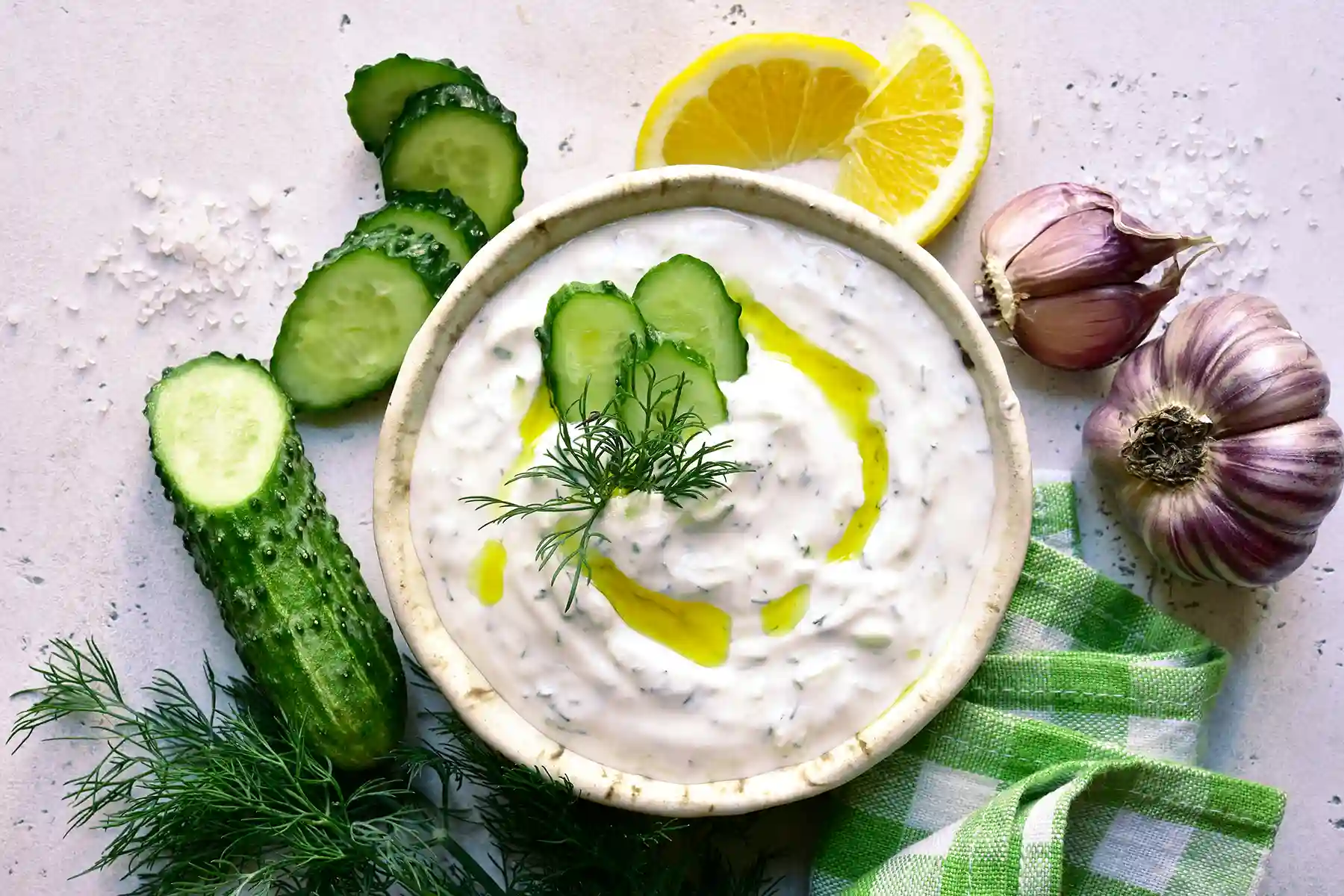 Tzatziki dip in a bowl with olive oil and dill with cucumbers around the bowl.