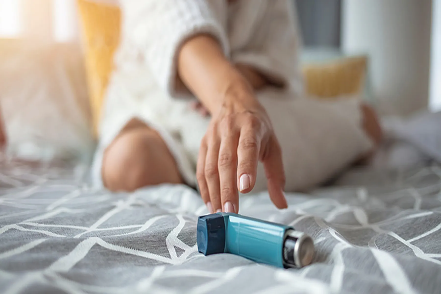 Woman sitting on bed reaches her hand out towards her blue inhaler