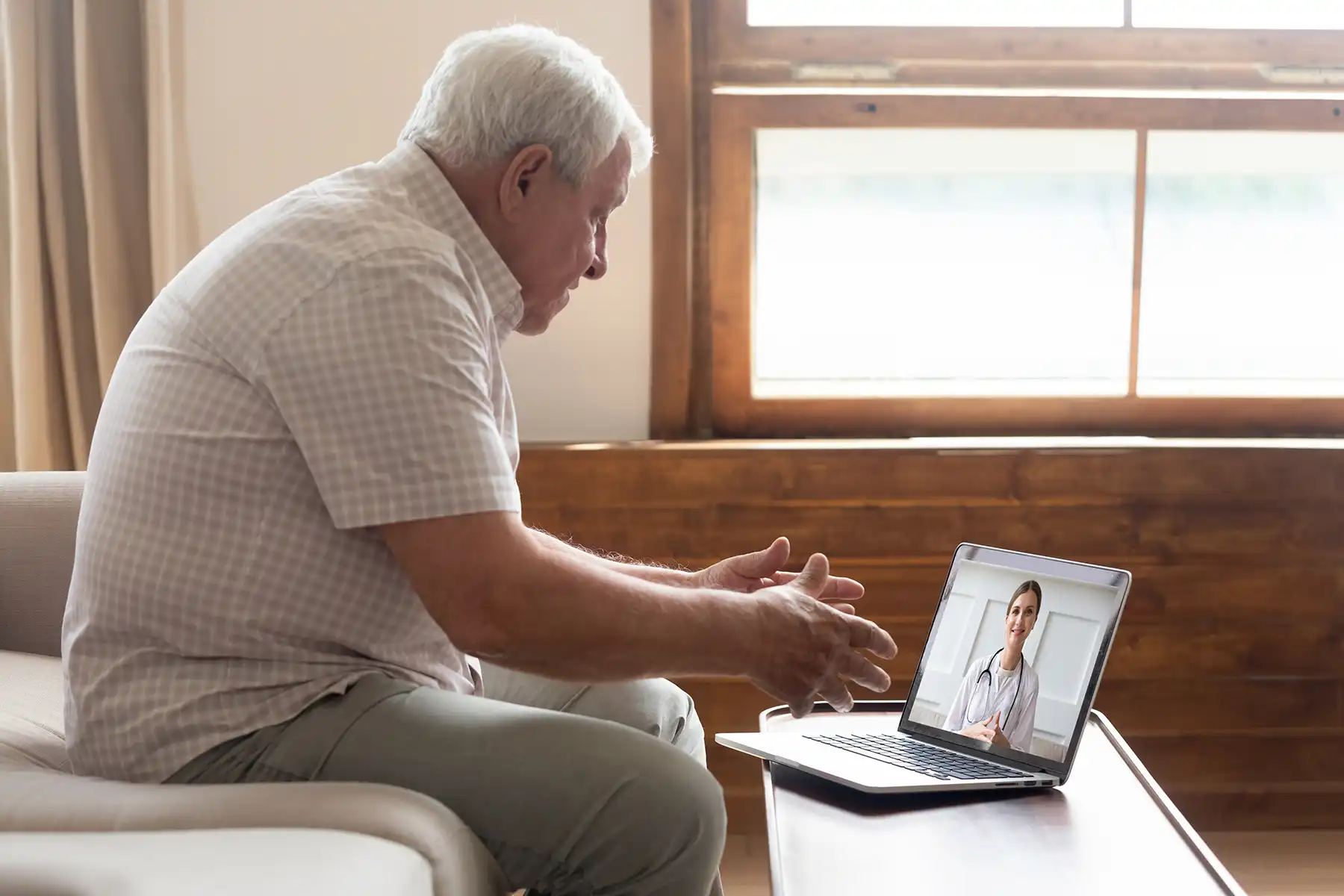 An older man sits on his couch while talking to his doctor via telehealth video appointment.