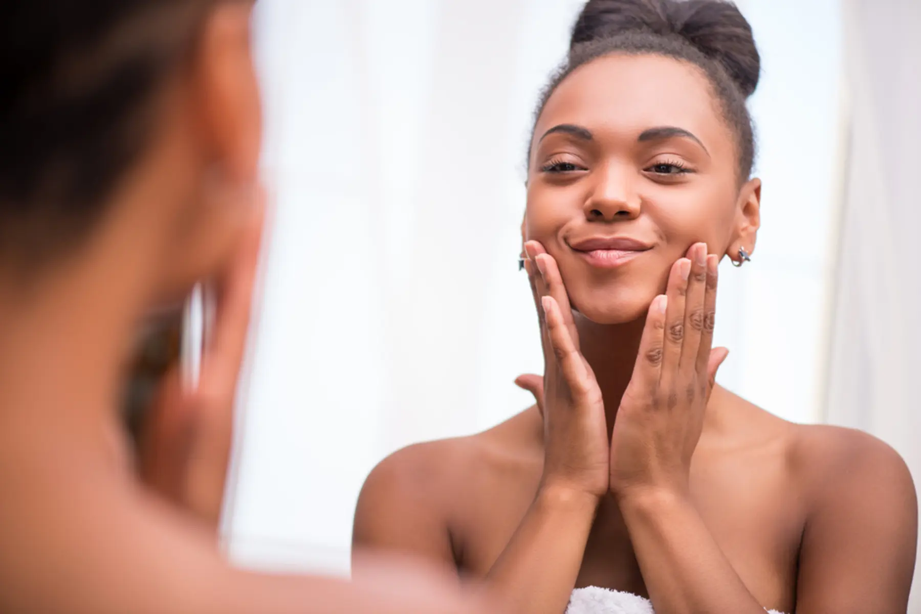 A woman looks in a mirror and applies skin care products.