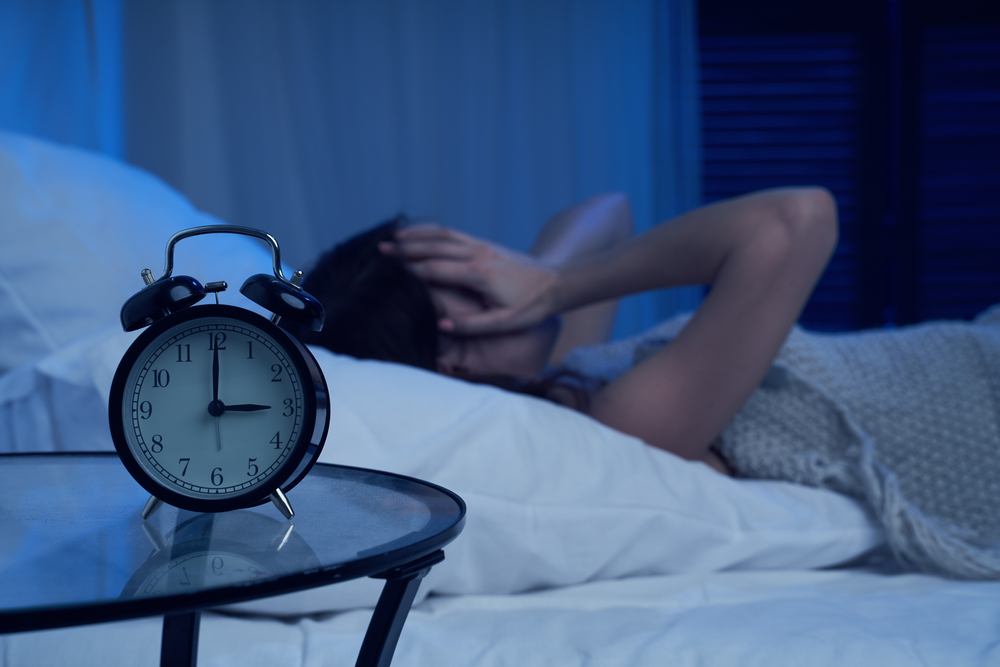 A woman lies in bed in a dark room with her hands on her face in frustration. There is a clock on the nightstand that says 3:00.