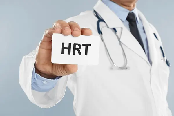 A doctor in a lab coat with a stethoscope around his neck holding a card toward the camera that says HRT. This stands for hormone replacement therapy. 