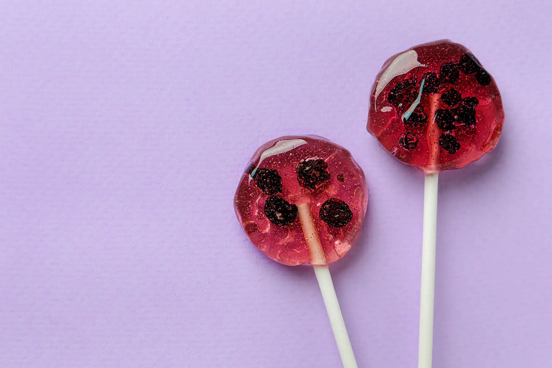 Sweet colorful lollipops with berries on lilac background, laying flat.