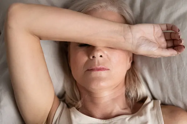 A woman lying on her back in bed or on a couch. Her arm is covering her eyes.