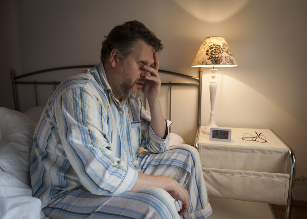 A man in blue, white, and yellow striped pajamas sits up on the side of the bed. He has his left hand on his face and is looking tired, frustrated, and/or upset. There is a lamp on at the nightstand beside him. 