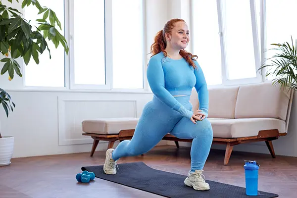 A woman in workout clothes does lunges in her living room with workout equipment around her.