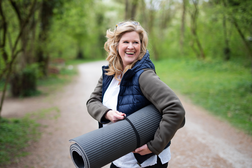 A middle-aged woman smiles as she holds a yoga or fitness mat outside. 