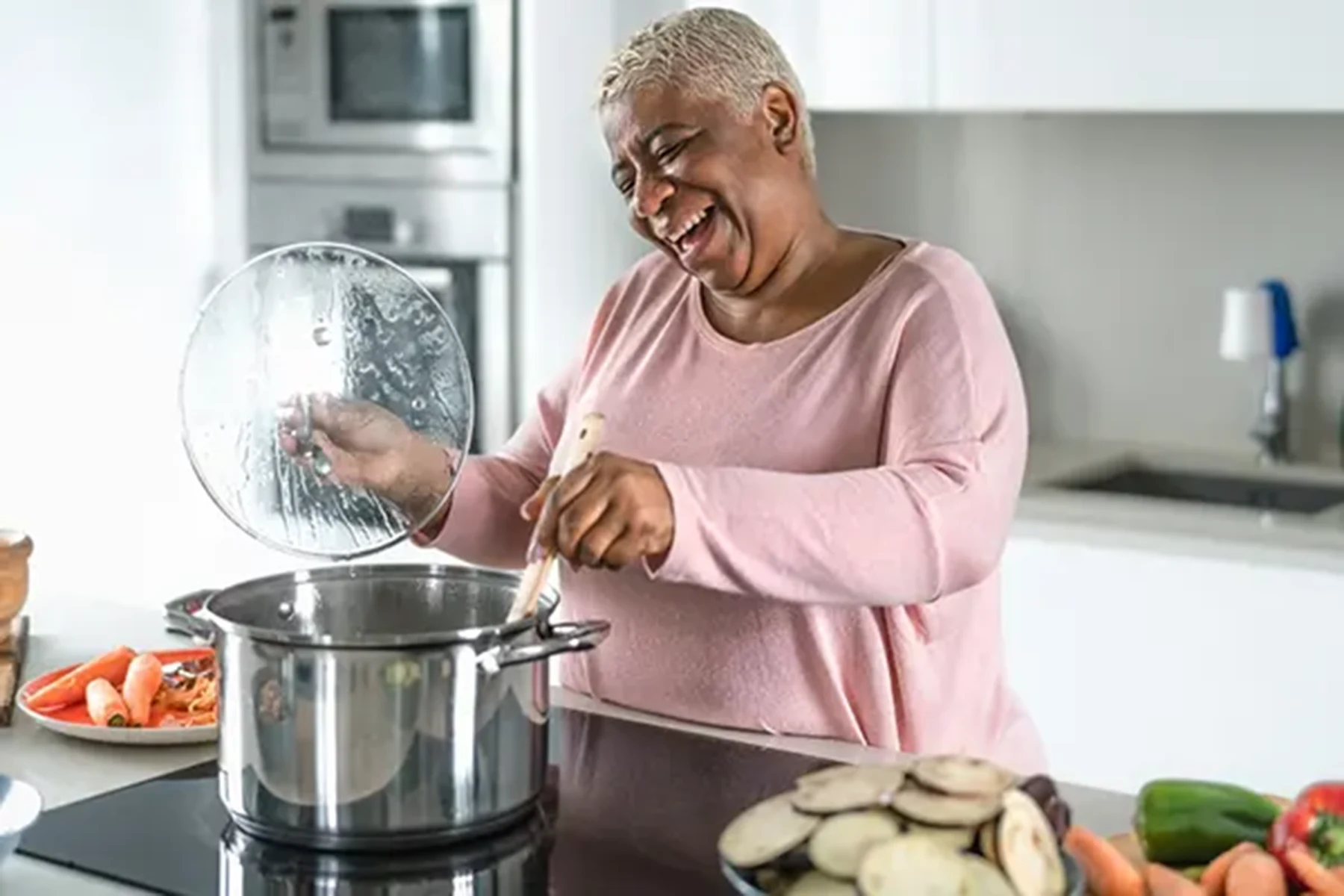 A woman stands at her stove with a pot and mixes, while smiling.