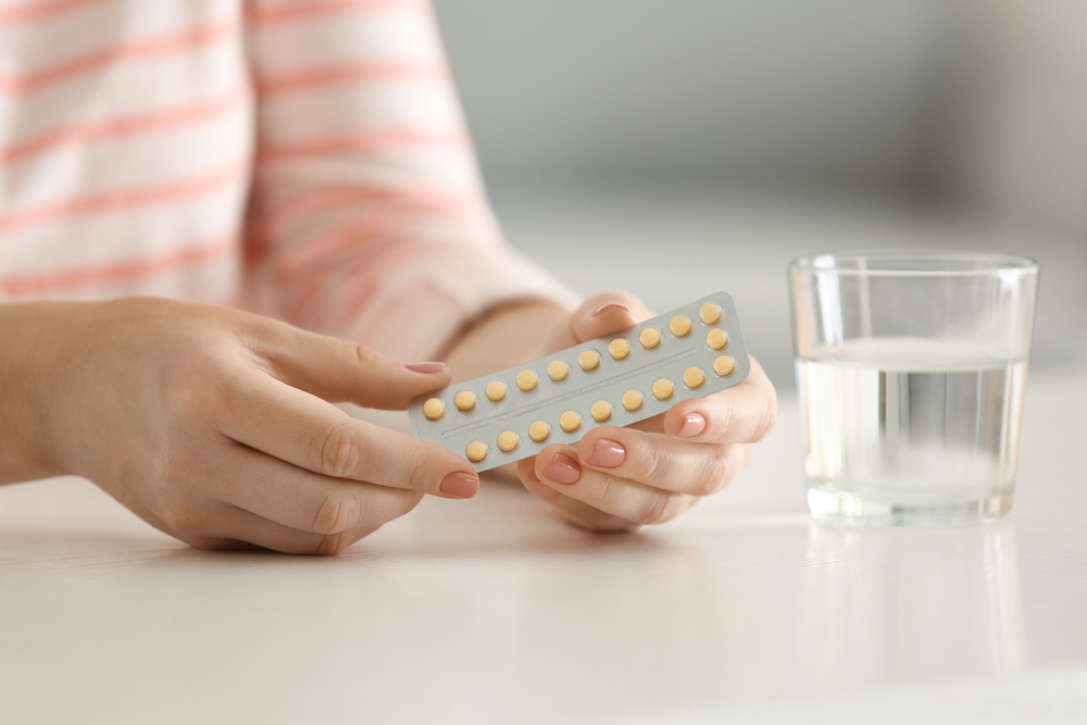 A woman holds a pack of birth control pills. A glass of water sits next to her on the table. 