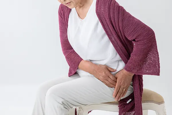 A woman in white shirt and pinkish/purplish sweater cardigan sits on a stool as she holds both her hands to her left hip in discomfort.