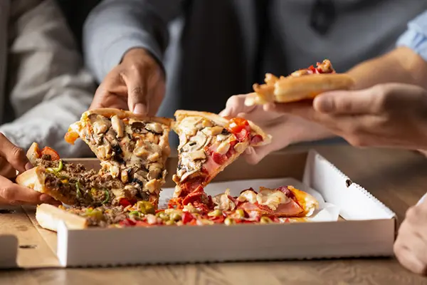 An open pizza box sits on a table as people grab slices from the box. The slices have a variety of meats and vegetables on them as toppings. 