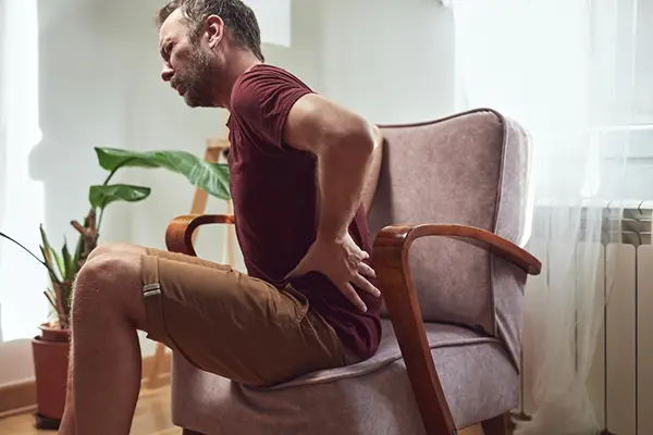 A man is sitting on a chair and holding his hip making a pained face.