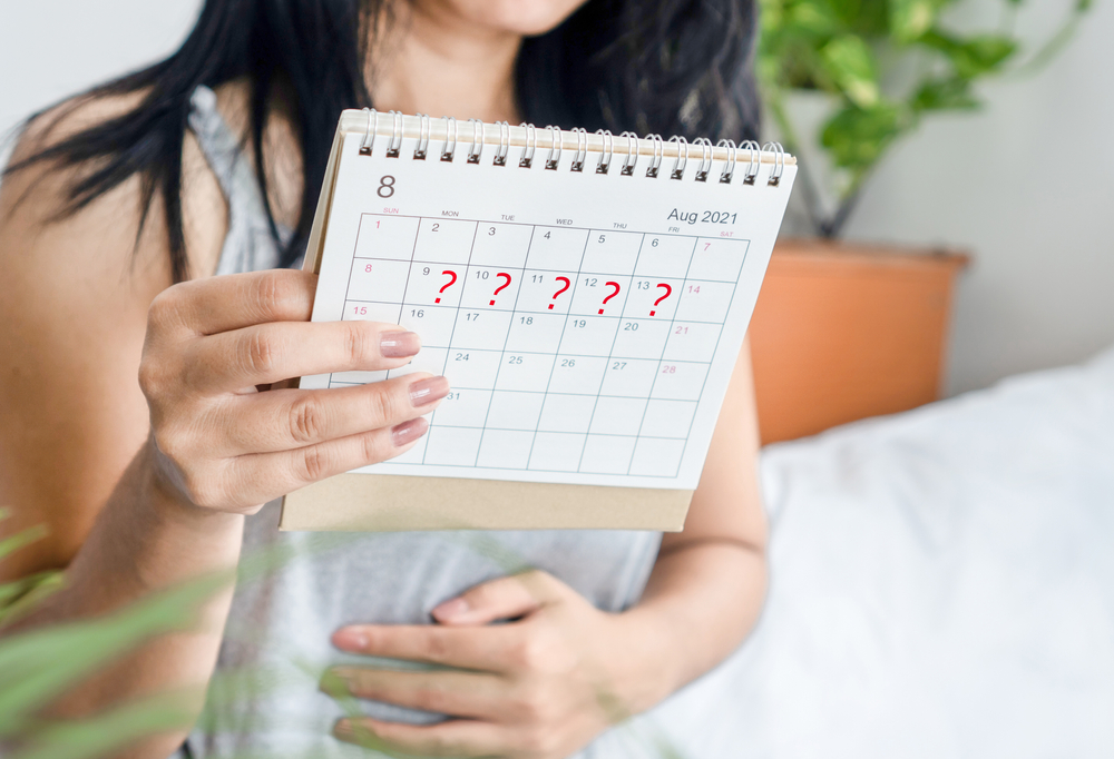 A woman with long dark hair holds up a calendar with question marks across five days of one week. Her other hand holds her stomach. 
