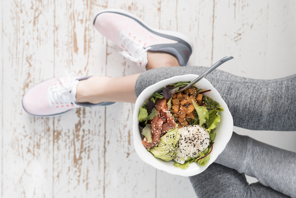 A top view of a woman sitting on the floor in workout clothes eating a salad filled with nutrient-rich ingredients.