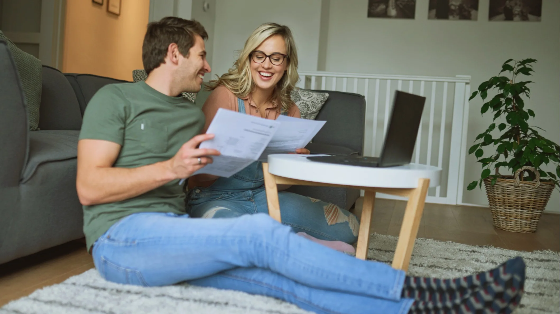 A smiling couple sitting on the floor, looking between paper documents and their laptop screen.