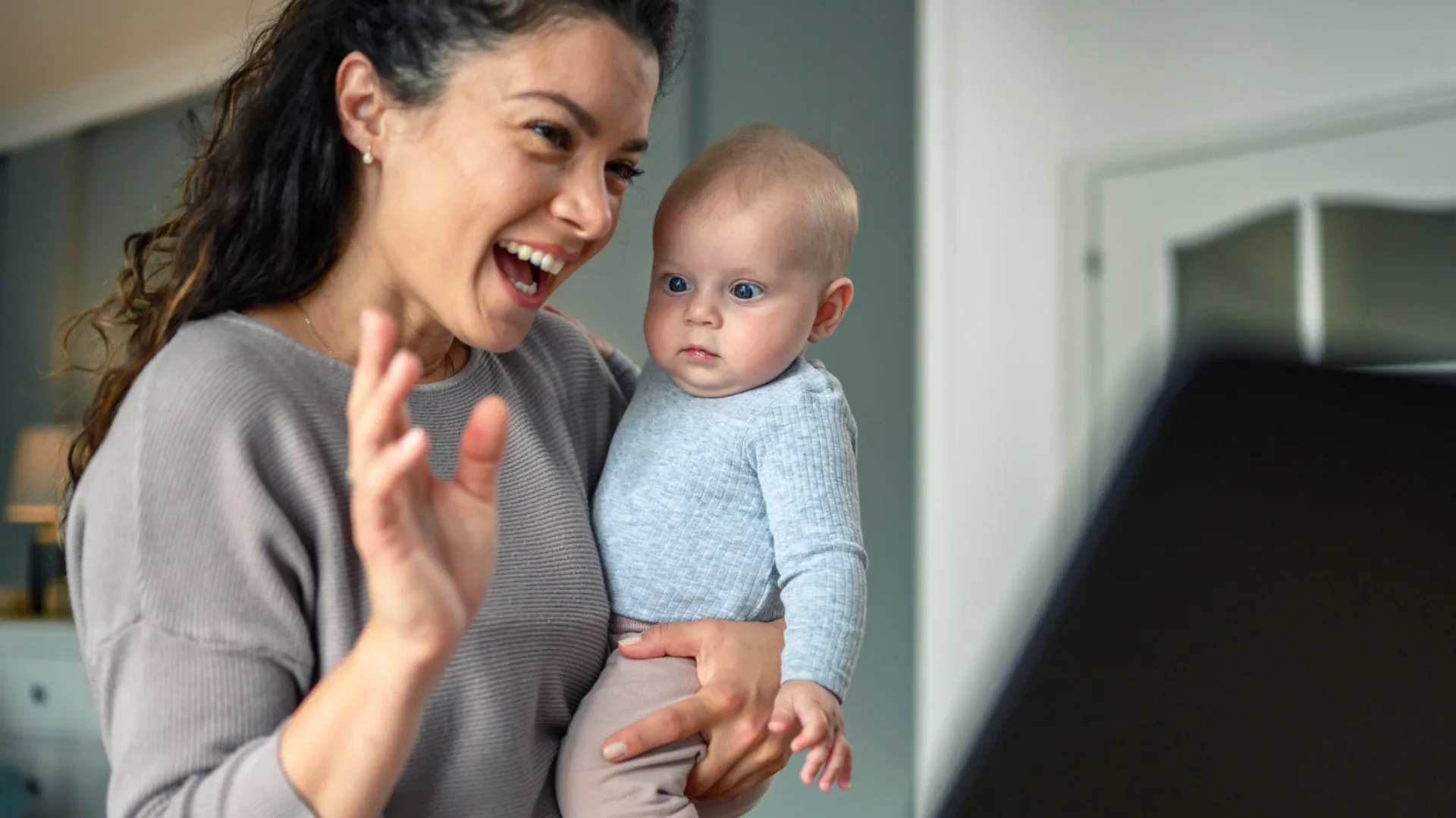 A woman holding a baby waving at a computer video call