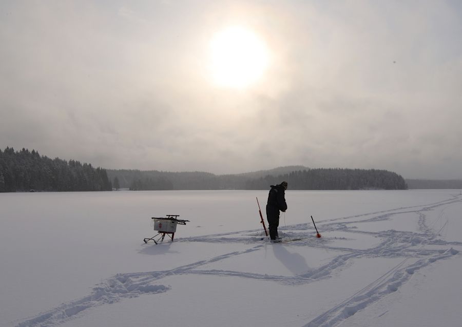 Kalastaja lumen peittämän järven päällä auringon paistaessa taivaalla pilvien takana
