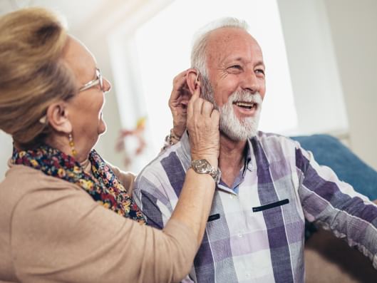 Die Seniorin hilft dem Senior, ein Hörgerät anzulegen.