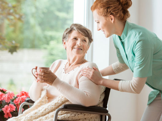 Pflegekraft mit Seniorin am Fenster.
