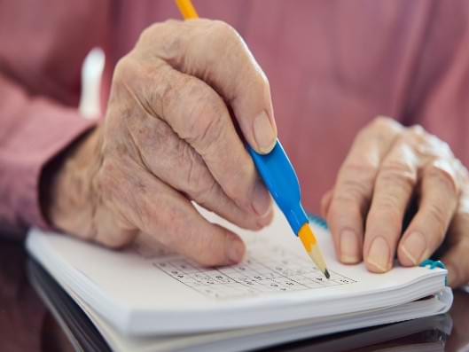 Ältere Person macht mit einem Bleistift mit Griffverdickung ein Rätsel.