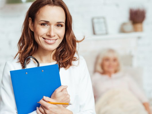 Eine Pflegerin mit blauem Klemmbrett. Im Hintergrund liegt eine Seniorin im Bett.