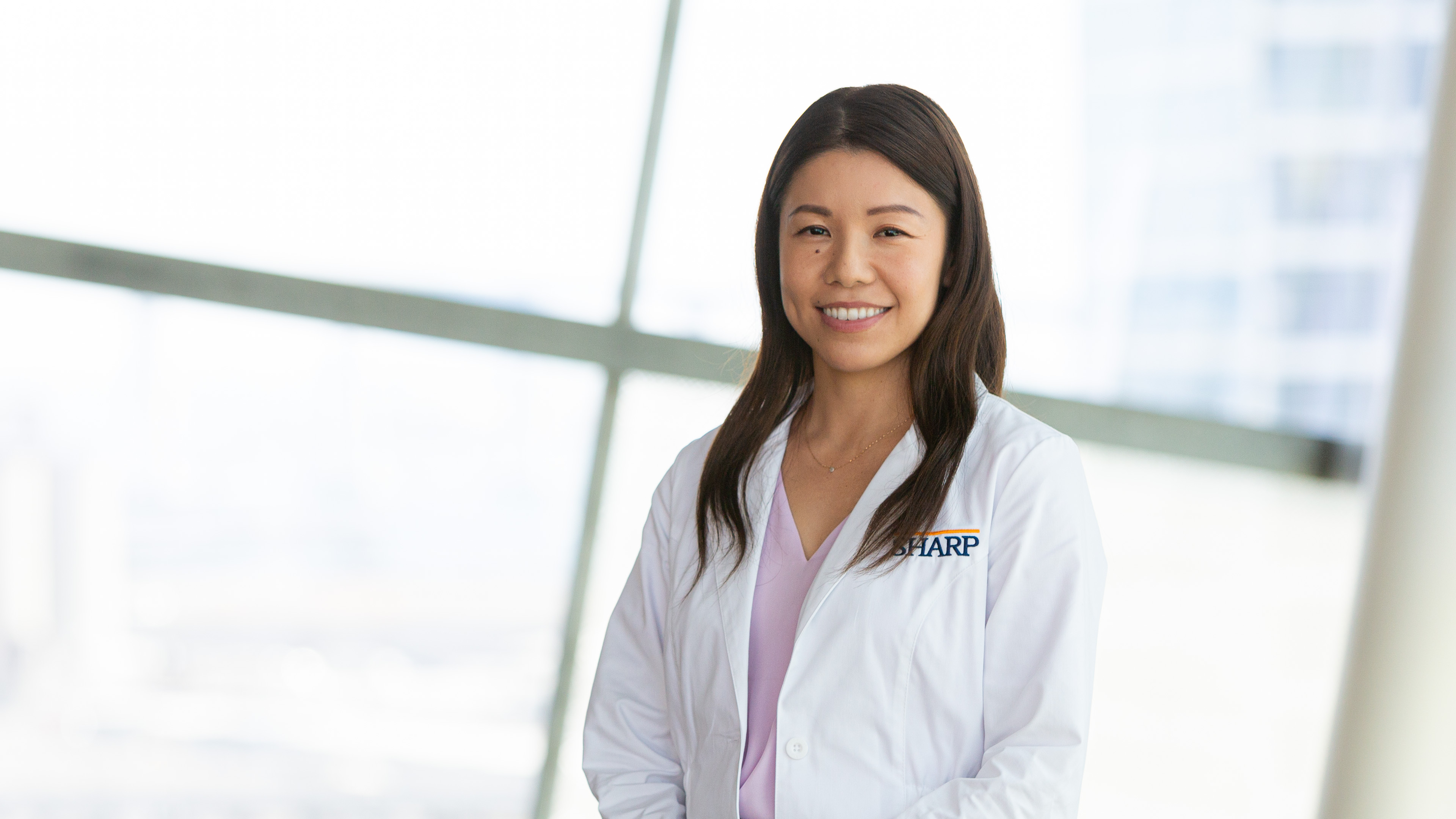 Dr. Nishimura, in a crisp white lab coat, offers a warm, inviting smile against a backdrop of sunlit windows, symbolizing a bright start to your healthcare journey with us