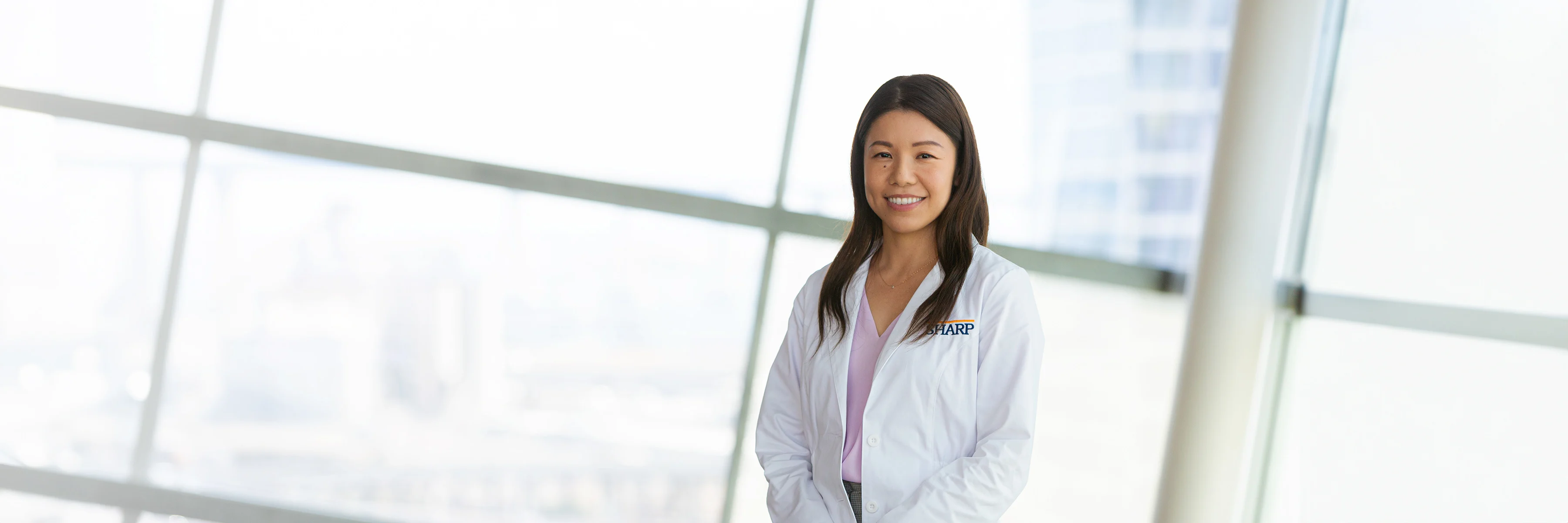 Dr. Nishimura, in a crisp white lab coat, offers a warm, inviting smile against a backdrop of sunlit windows, symbolizing a bright start to your healthcare journey with us