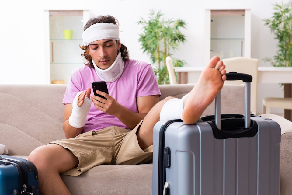 Mann mit verschiedenen Verbänden sitzt auf dem Sofa und kontrolliert die Deckung seiner Grundversicherung im Ausland. Den Fuss lagert er auf einem Koffer hoch.