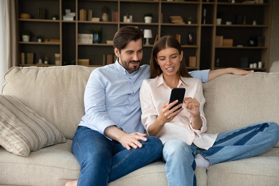 Mann und Frau sitzen auf der Couch, sie schauen gemeinsam auf ein Handy und erkundigen sich über die Voraussetzungen eines Kredits.